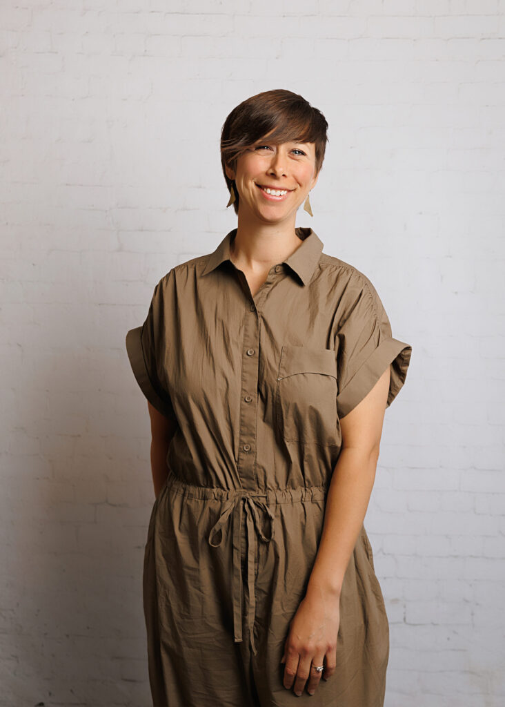 Abby Simon, artist and co-founder of Simon Maker Studio, standing against a white brick wall wearing a tan dress and handmade earrings, smiling warmly.