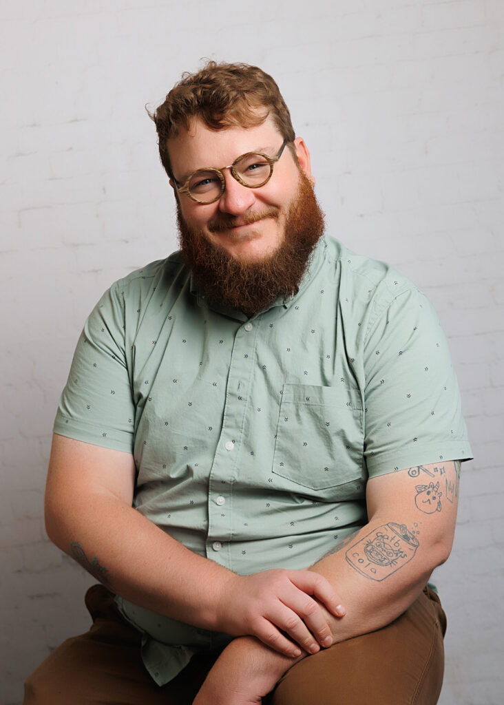 Peter Simon, co-founder of Simon Maker Studio in Viroqua, Wisconsin, smiling with arms crossed in front of a white brick wall. He is wearing glasses and a light green shirt with small printed arrows