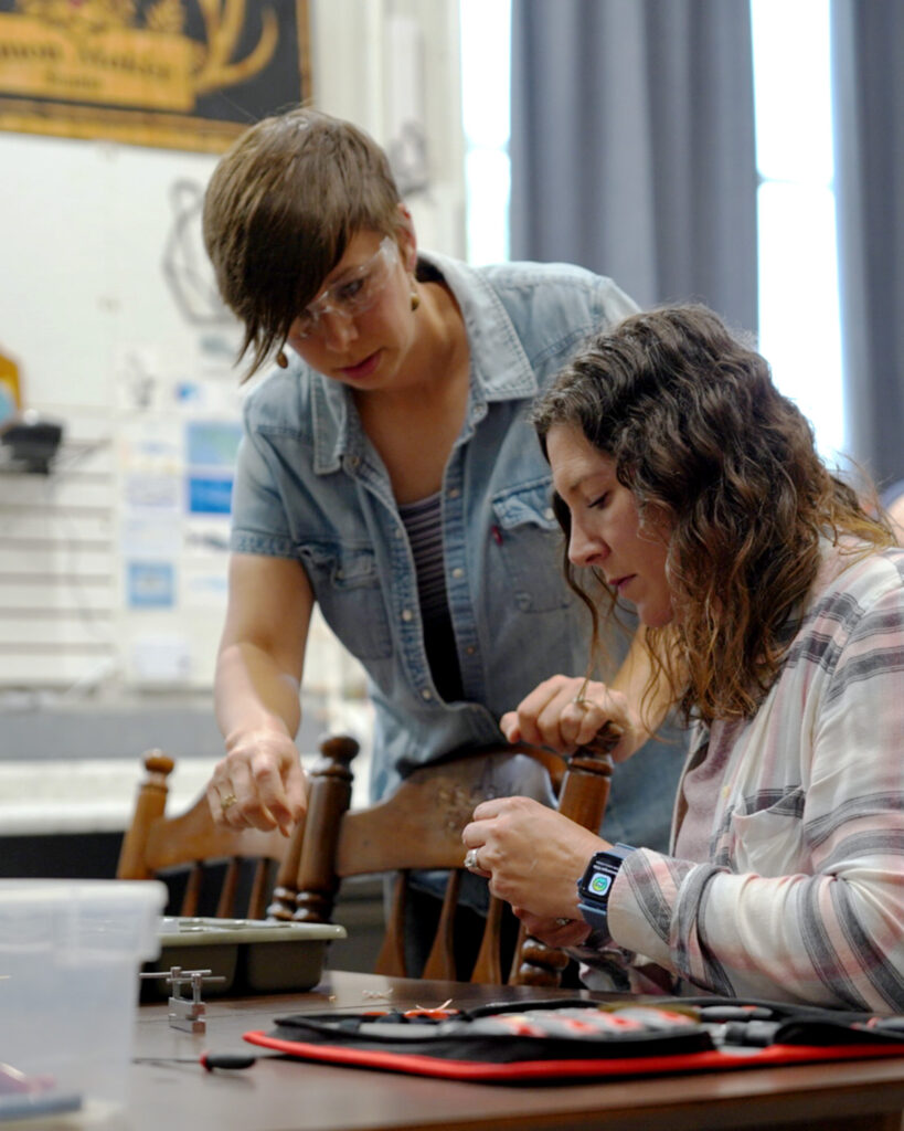Abby Simon of Simon Maker Studio in Viroqua, Wisconsin, helping a participant during a hands-on jewelry-making workshop. The artist provides guidance as the student works with copper wire at a shared studio table.